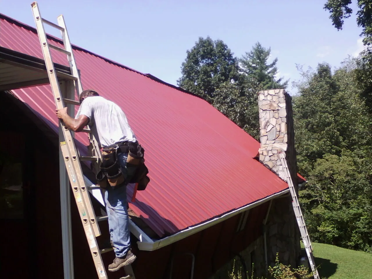 Roofer on a ladder beside a red metal roof and stone chimney — Soffit & Fascia Repair by Roofing Brothers in Horseshoe Bend